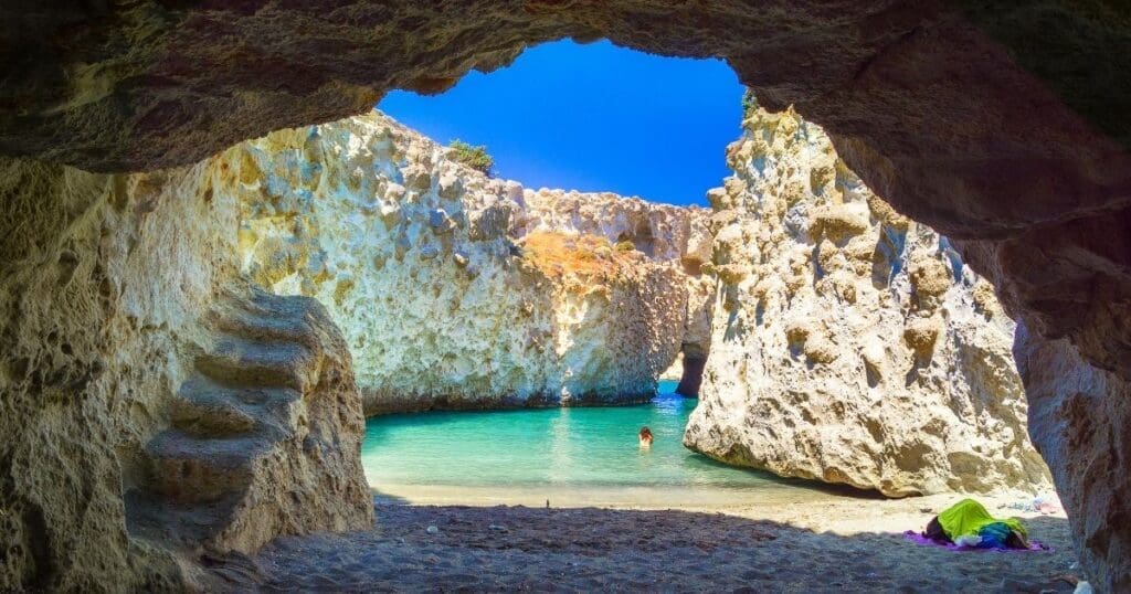 Looking out from inside a coastal stone sea cave at the bright turquoise water and rugged cliffs of Antiparos, Greece.