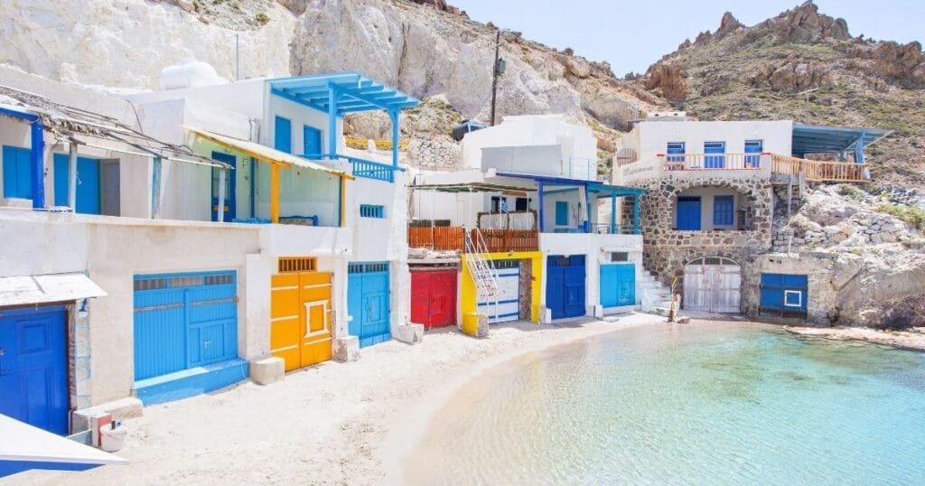 Traditional two-story fisherman houses with brightly painted yellow, blue, and red doors along the sandy waterfront of Klima village in Milos, Greece.